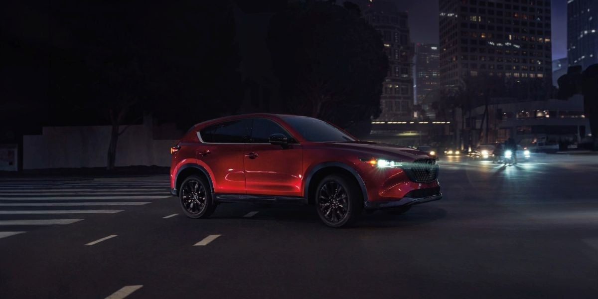 Red Mazda CX-5 driving through a city at night with illuminated buildings in the background.