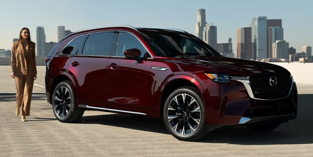 2025 Mazda CX-90 in a deep red color parked on a rooftop with a city skyline in the background, woman walking nearby.